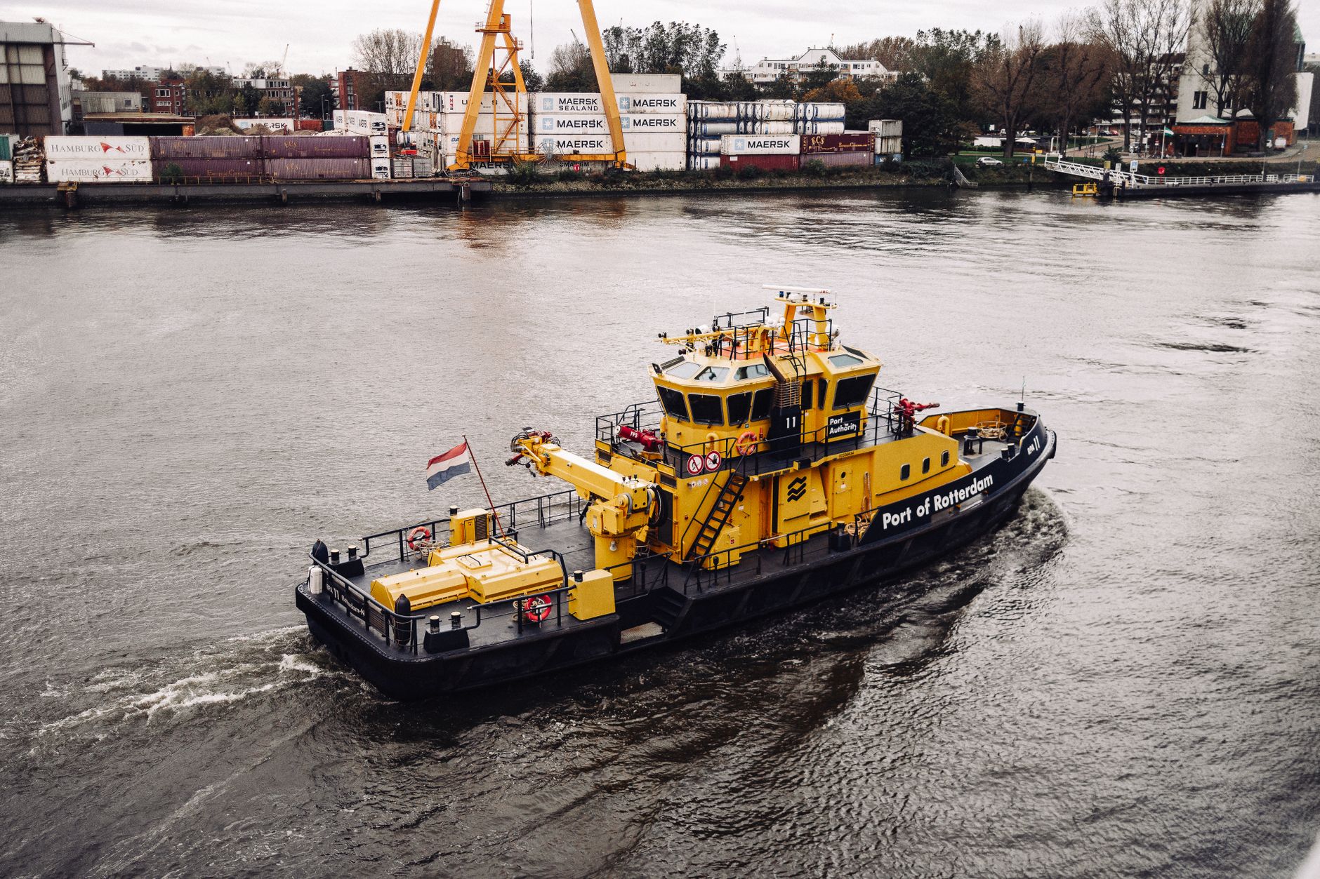 Een gele sleepboot vaart op de rivier in de haven van Rotterdam, met containers en een kraan op de achtergrond.