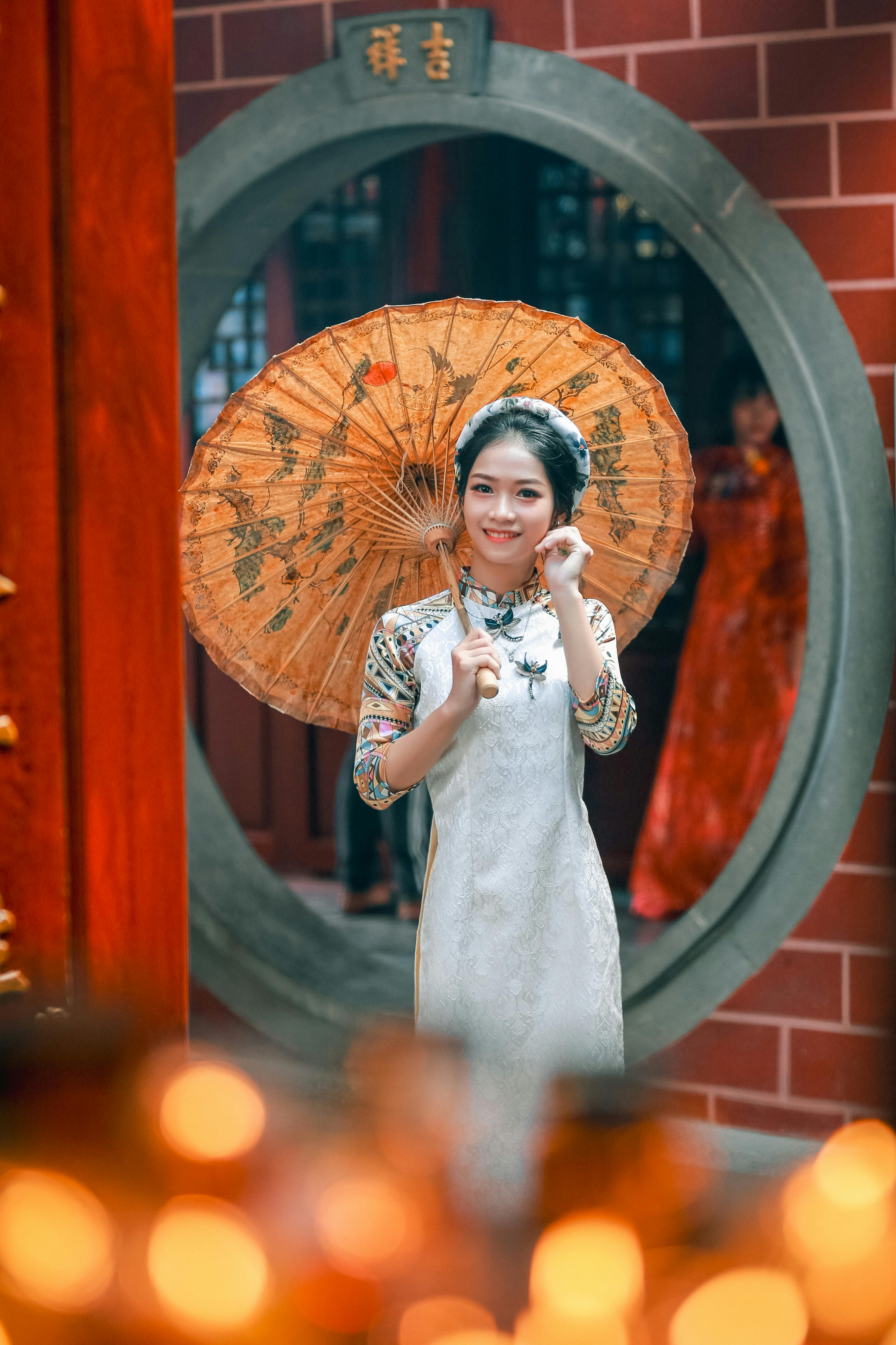 A woman in ao dai with a paper parasol stands in a traditional Vietnamese setting.