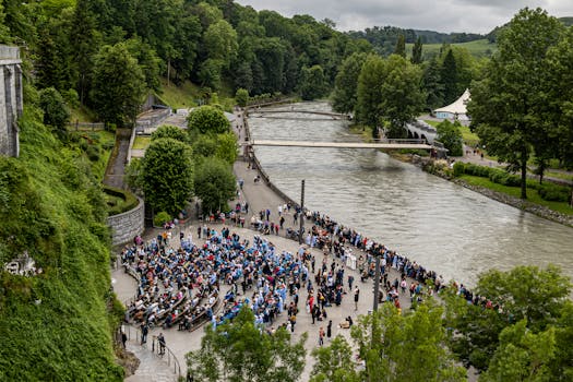 A large crowd gathers by a scenic riverside park surrounded by lush greenery on a cloudy day.