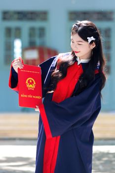 Young woman in cap and gown holding diploma outdoors on a sunny day.