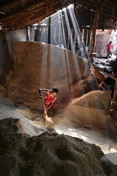 阳光明媚的仓库里，一名男子正在铲材料，光线从屋顶的缝隙中倾泻而下