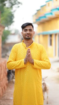 Young man wearing a yellow traditional outfit, expressing a greeting gesture outdoors in a rural setting.