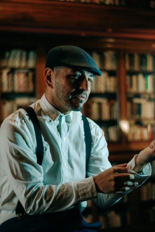 Free A stylish man in a cap sits in a vintage library, wearing suspenders and a white shirt. Stock Photo