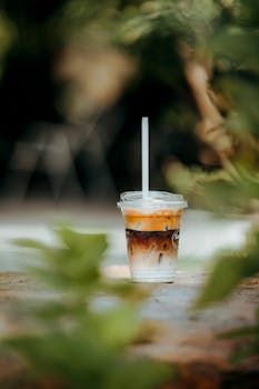 A single iced coffee cup with a straw in a lush outdoor setting, surrounded by blurred greenery.