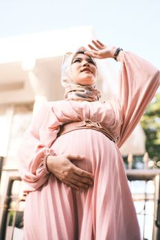 Pregnant woman in a pink dress and hijab standing outdoors, hand on belly. - Fotografer Sidoarjo