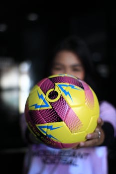 A vibrant yellow and pink soccer ball is held up, obscuring a person's face.