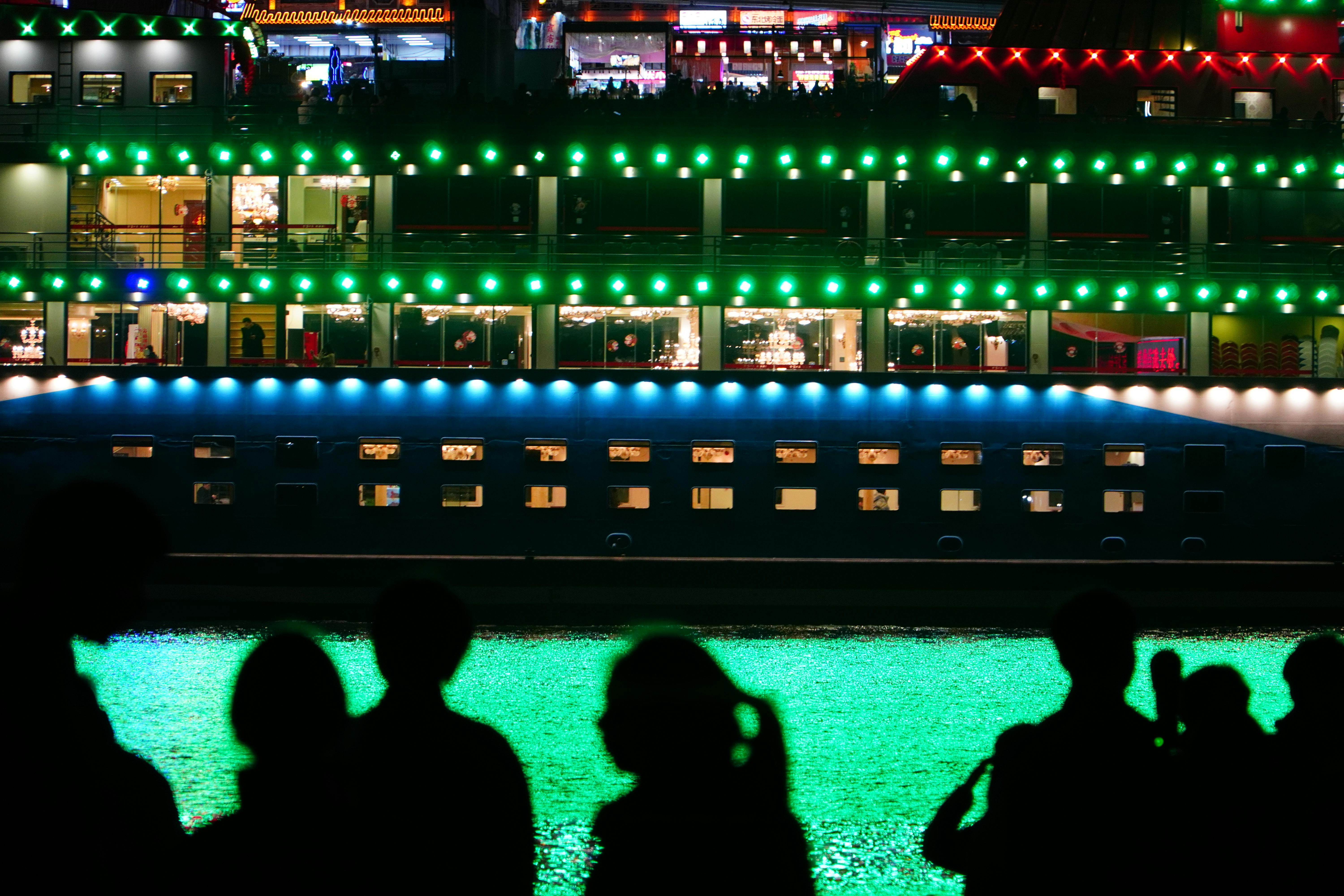 Silhouettes observing a brightly lit cruise ship at night, a lively waterside scene.