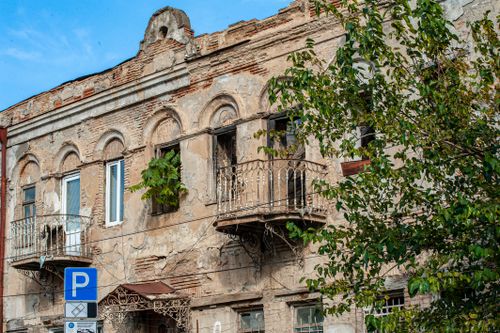 Free A weathered building with ornate balconies and a 'P' parking sign, partially obscured by leafy trees. Stock Photo
