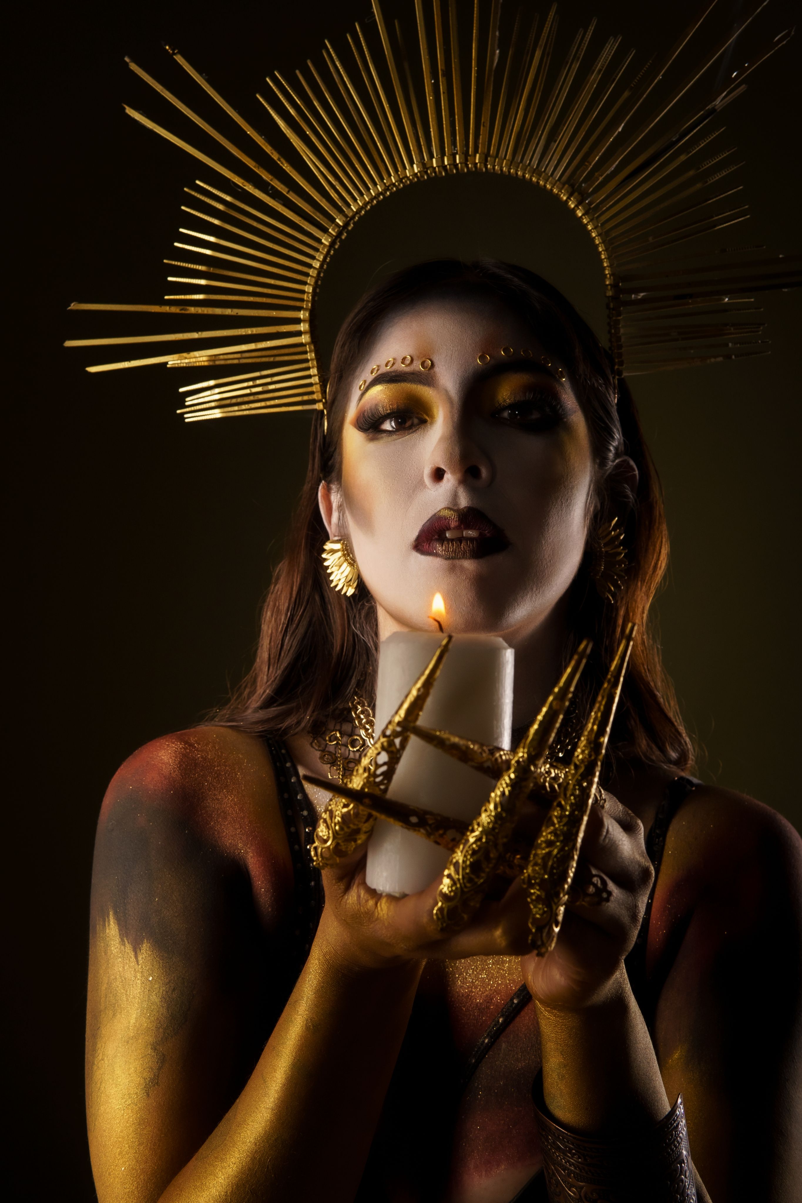 A dramatic portrait of a woman wearing gold accessories, holding a candle, with striking makeup and halo.
