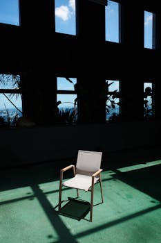 A lone chair sits in a shadowy indoor space with window reflections.