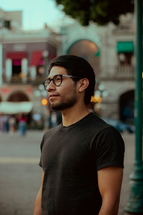 Free A young man with glasses gazes thoughtfully in a lively urban setting. Stock Photo