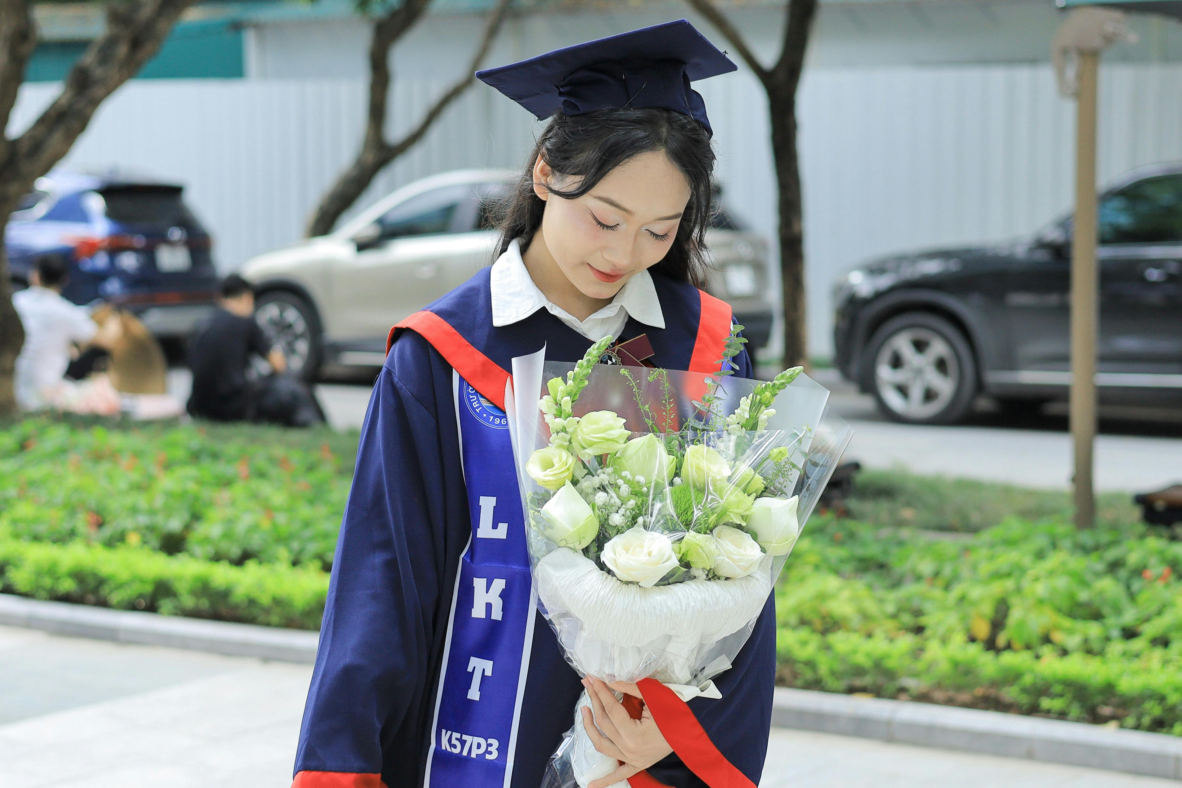 Young woman in graduation attire holding a bouquet of white flowers outdoors in Việt Nam.