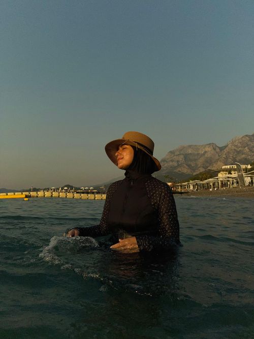 Free Woman wearing hat and hijab stands in ocean water near picturesque beach with mountains. Stock Photo