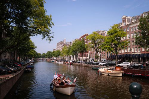 Free Tourists enjoying a sunny boat ride on a picturesque Amsterdam canal lined with historic buildings. Stock Photo