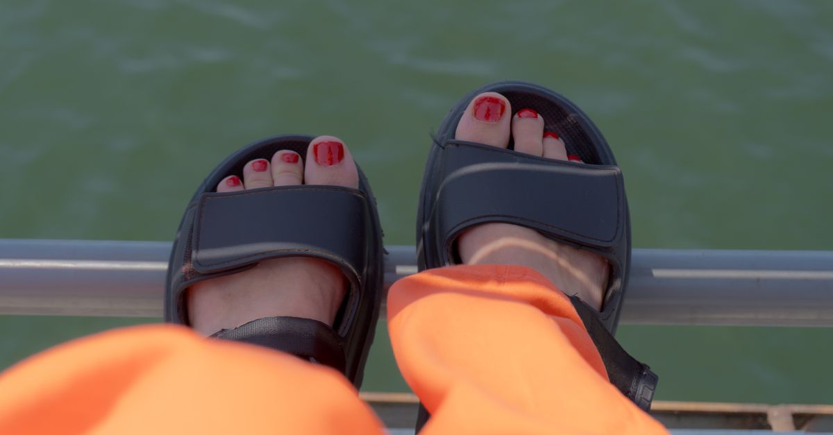 Feet resting on a railing above water, wearing black sandals and orange pants.