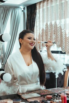 Woman in a salon applying makeup while wearing a chic white robe, preparing for an event.