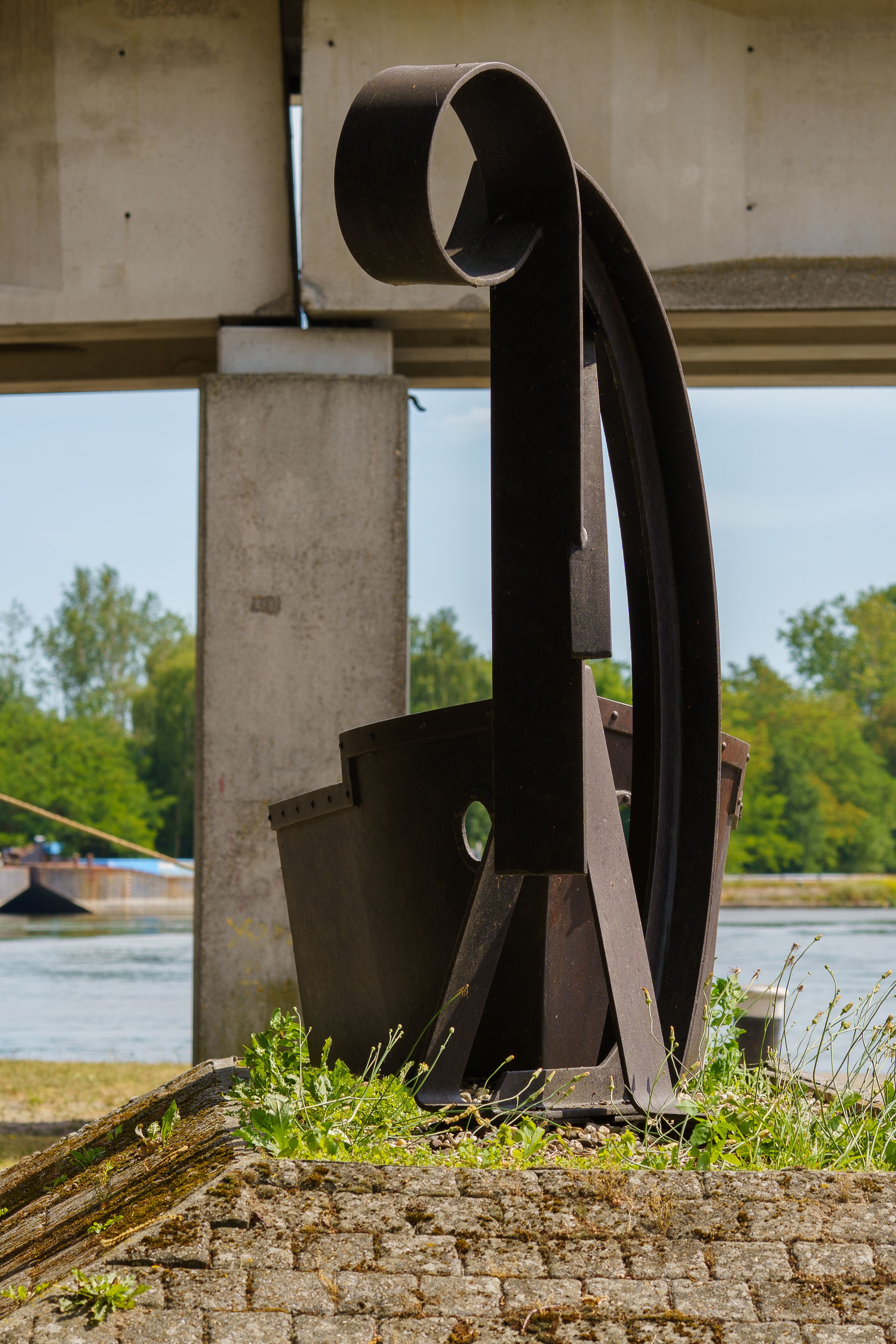 Artistic metal sculpture by a canal in Dessel, Belgium, under a bridge.