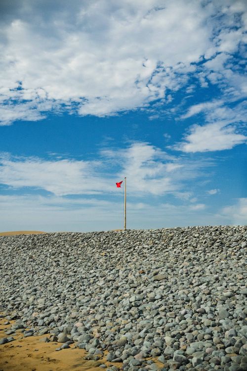 Free A rocky beach in Maspalomas with a red warning flag against a vibrant blue sky and scattered clouds. Stock Photo