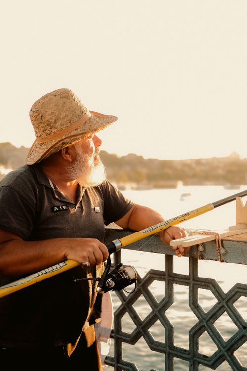 Free Senior man with fishing rod enjoying sunset by the waterfront, wearing a straw hat. Stock Photo