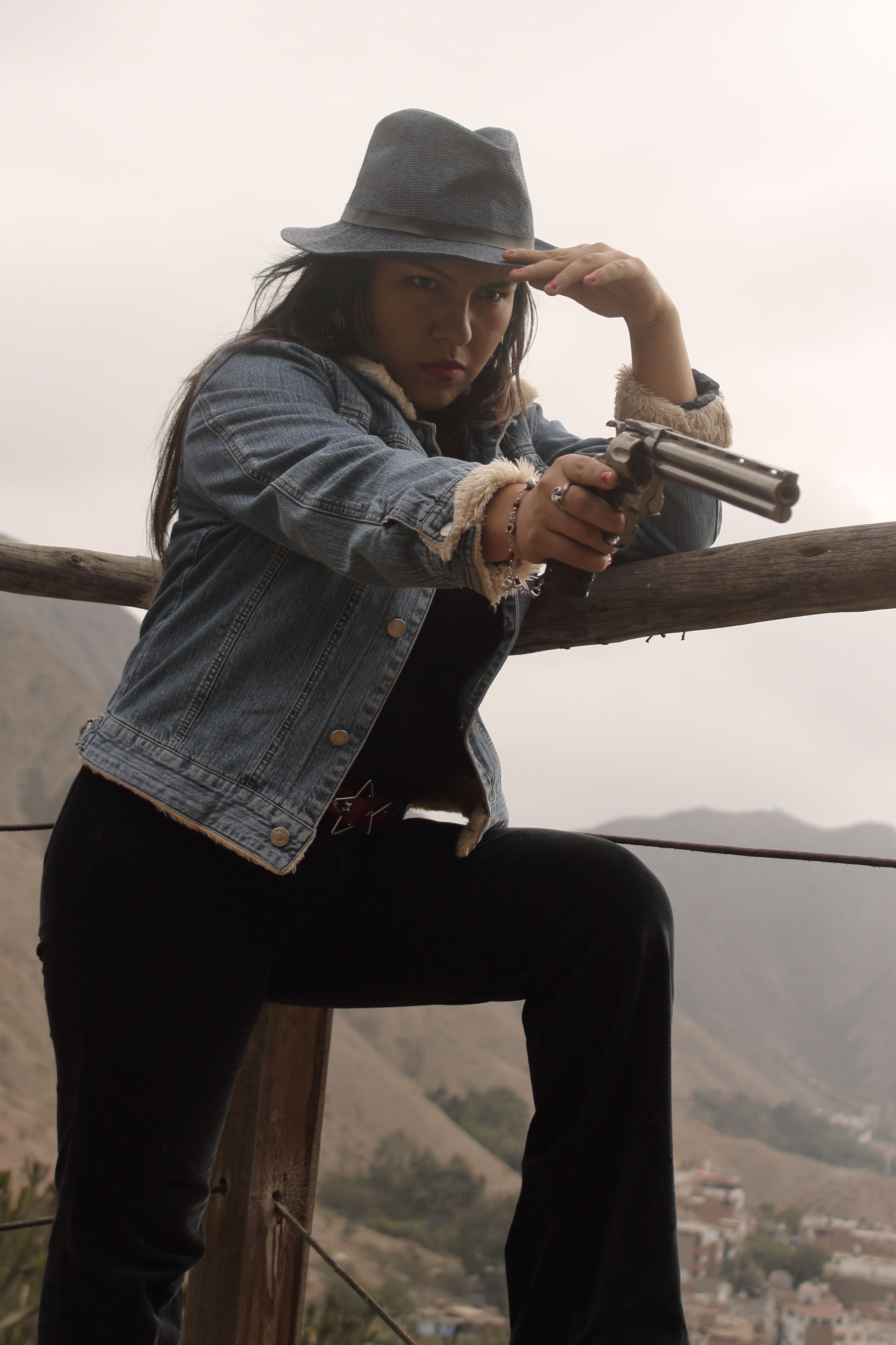 Woman in western attire holding a revolver, posing against a scenic mountain backdrop in Peru.