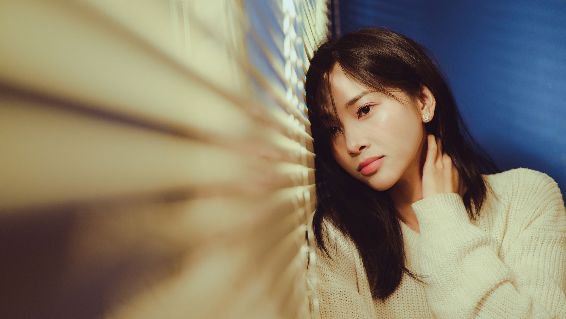 Thoughtful woman in cozy sweater gazing through window blinds.