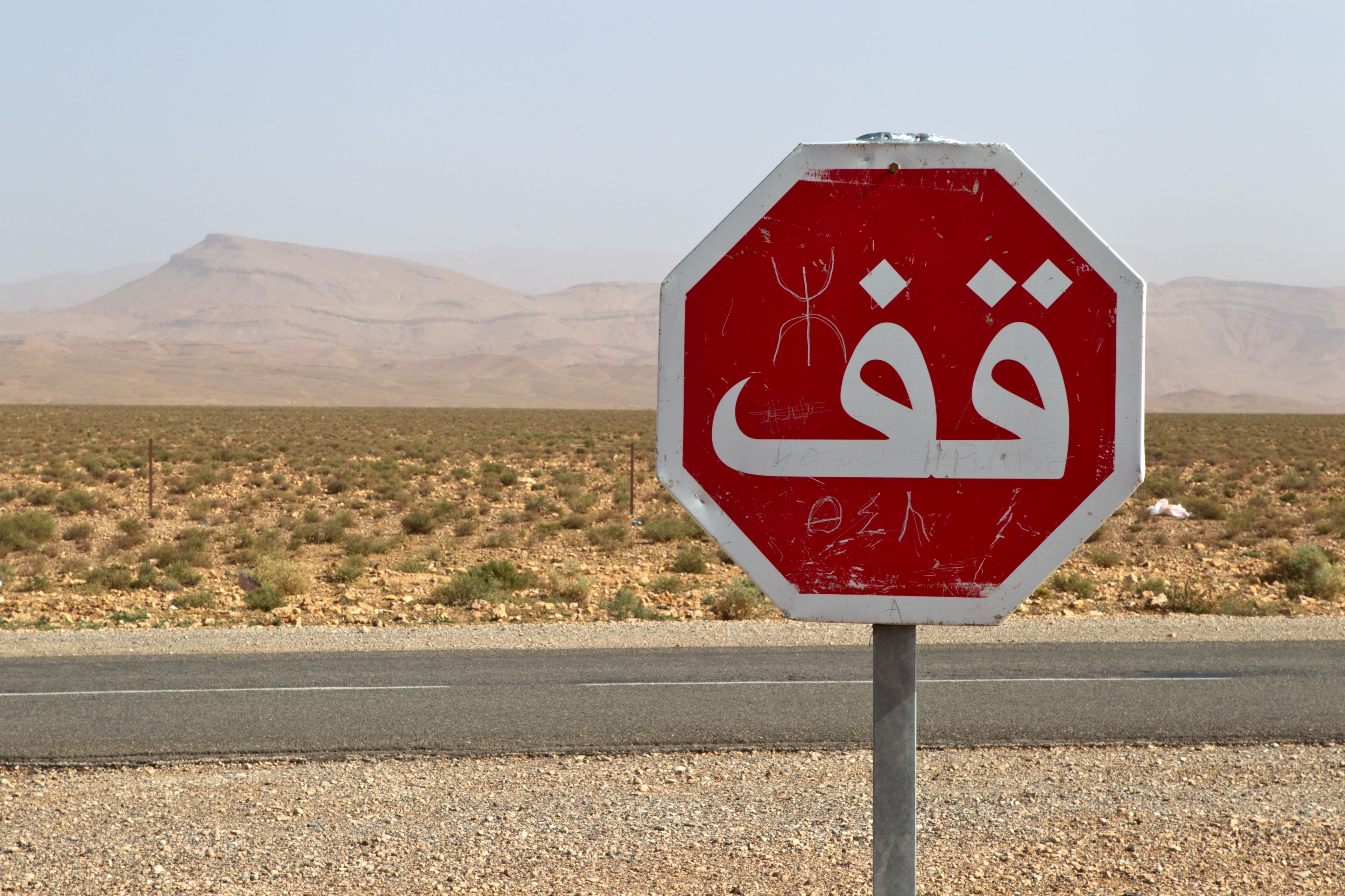 A red stop sign with Arabic writing stands in the vast Moroccan desert under a clear sky.
