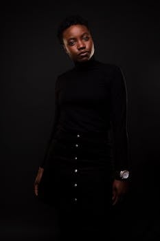 一位身着时尚黑衣的黑人女性，在暗色背景前优雅地摆出姿势
