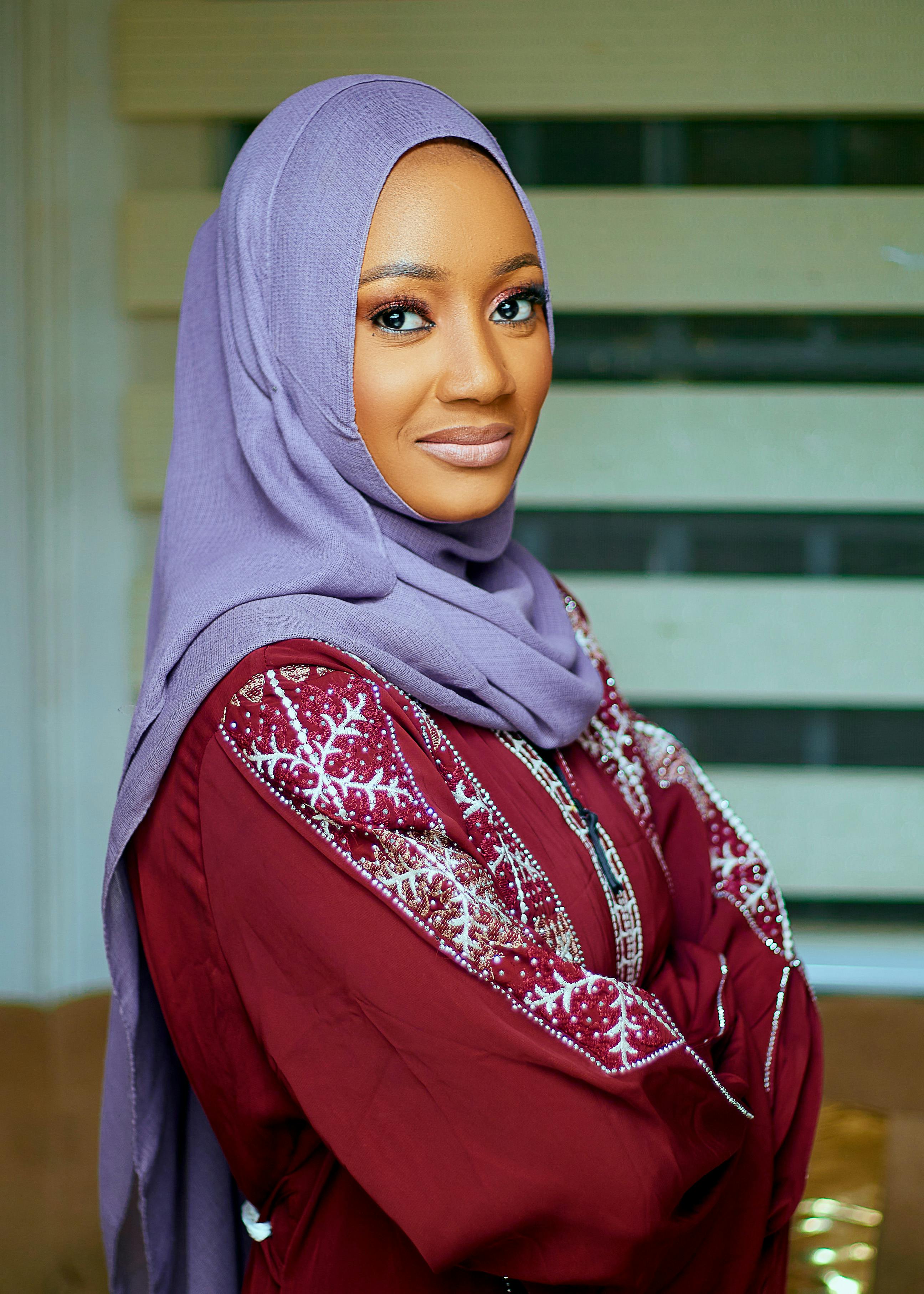 Vibrant portrait of smiling woman in lavender hijab and embroidered maroon dress.