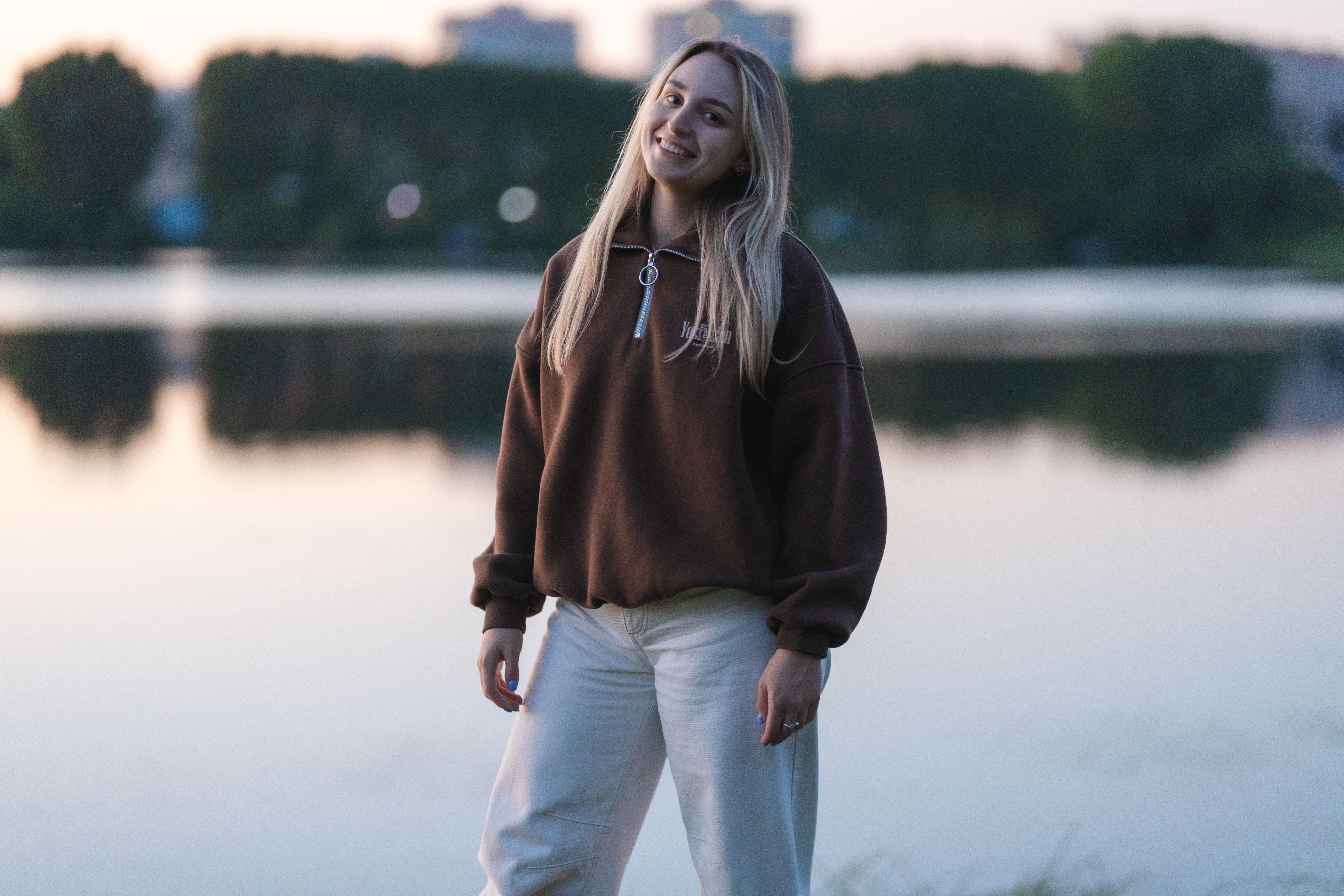 Femme souriante portant un pull quarter zip marron au bord d'un lac au crépuscule