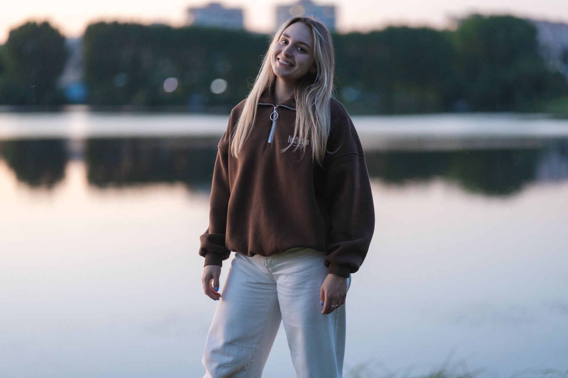 Femme souriante portant un pull quarter zip marron au bord d'un lac au crépuscule