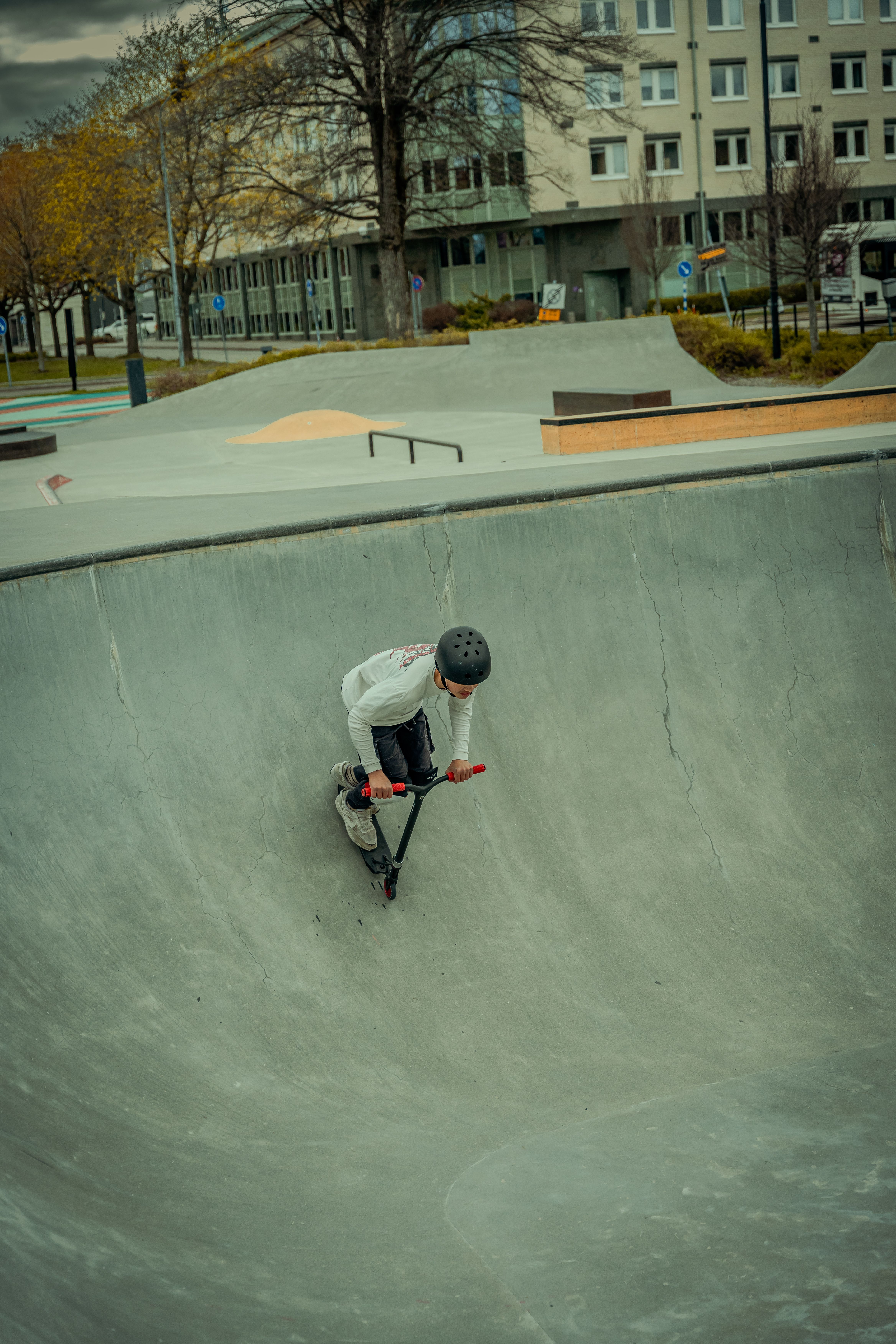 A young boy skillfully rides a scooter in an urban skatepark during a cool day.