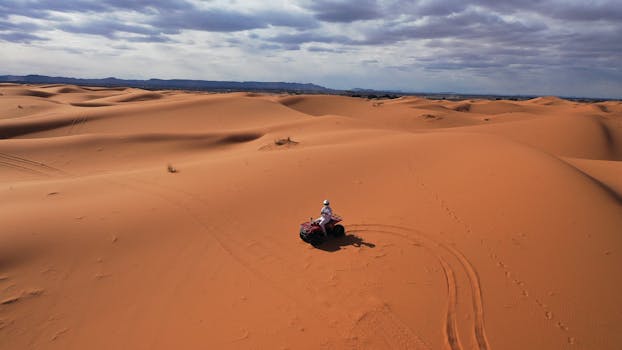 A quad biker navigates the expansive orange dunes of the Sahara Desert under a cloudy sky.