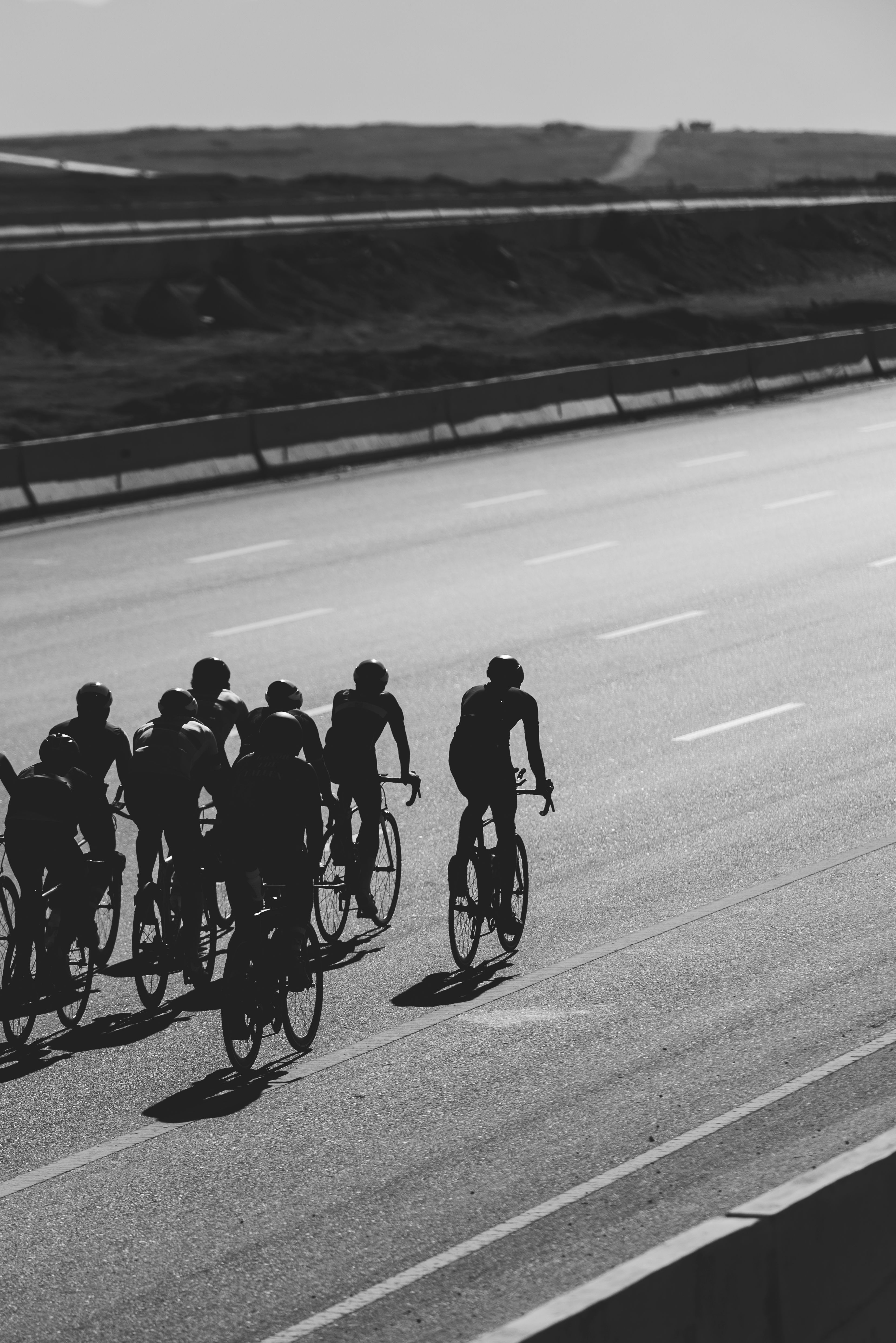 A group of cyclists rides on an empty highway under clear skies, captured in monochrome.