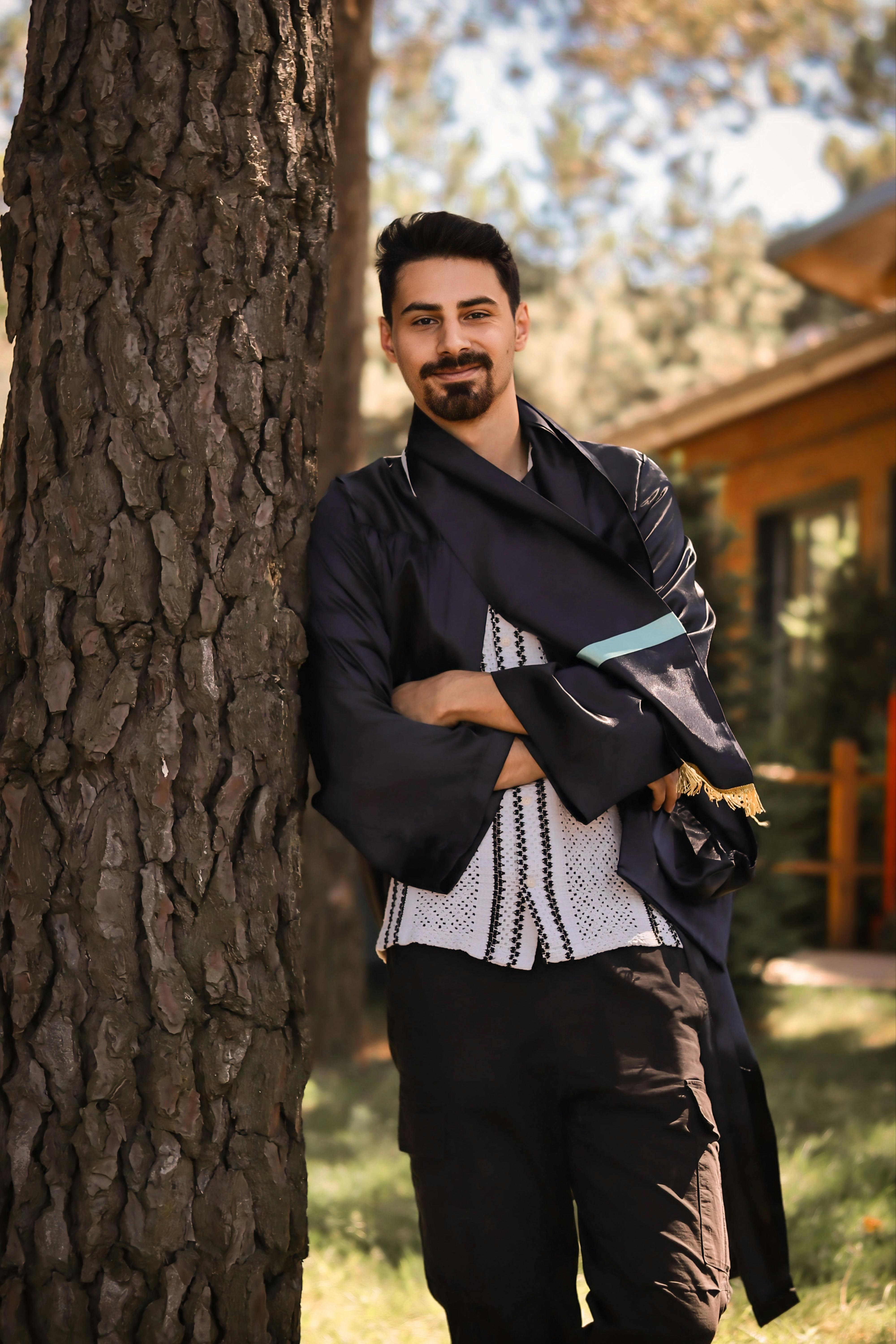 A smiling man in traditional clothing leans against a tree outdoors on a sunny day.