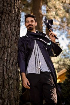 Portrait of a smiling young man in graduation attire, standing outdoors next to a tree. - Fotografer Sidoarjo