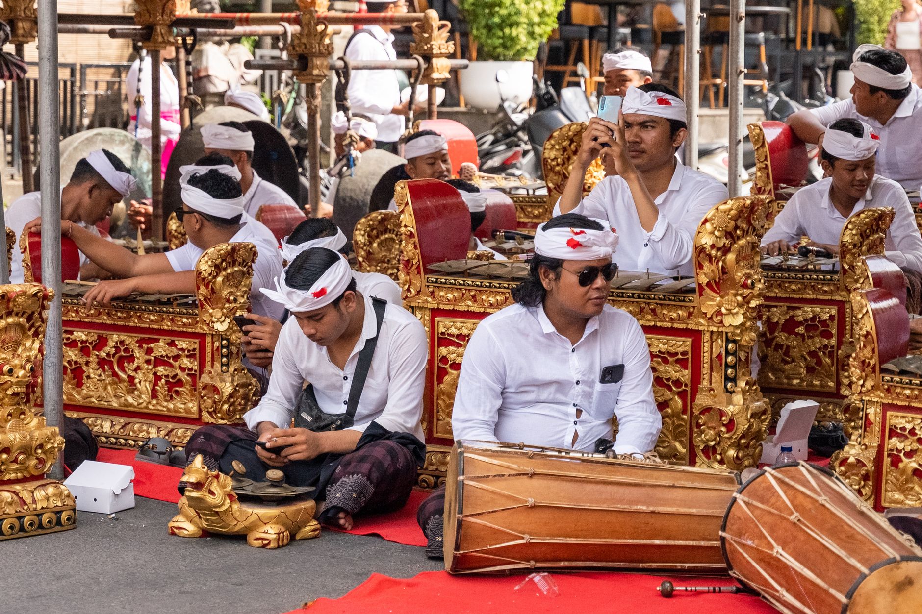 Balinese musicians perform traditional gamelan music outdoors, wearing cultural attire.