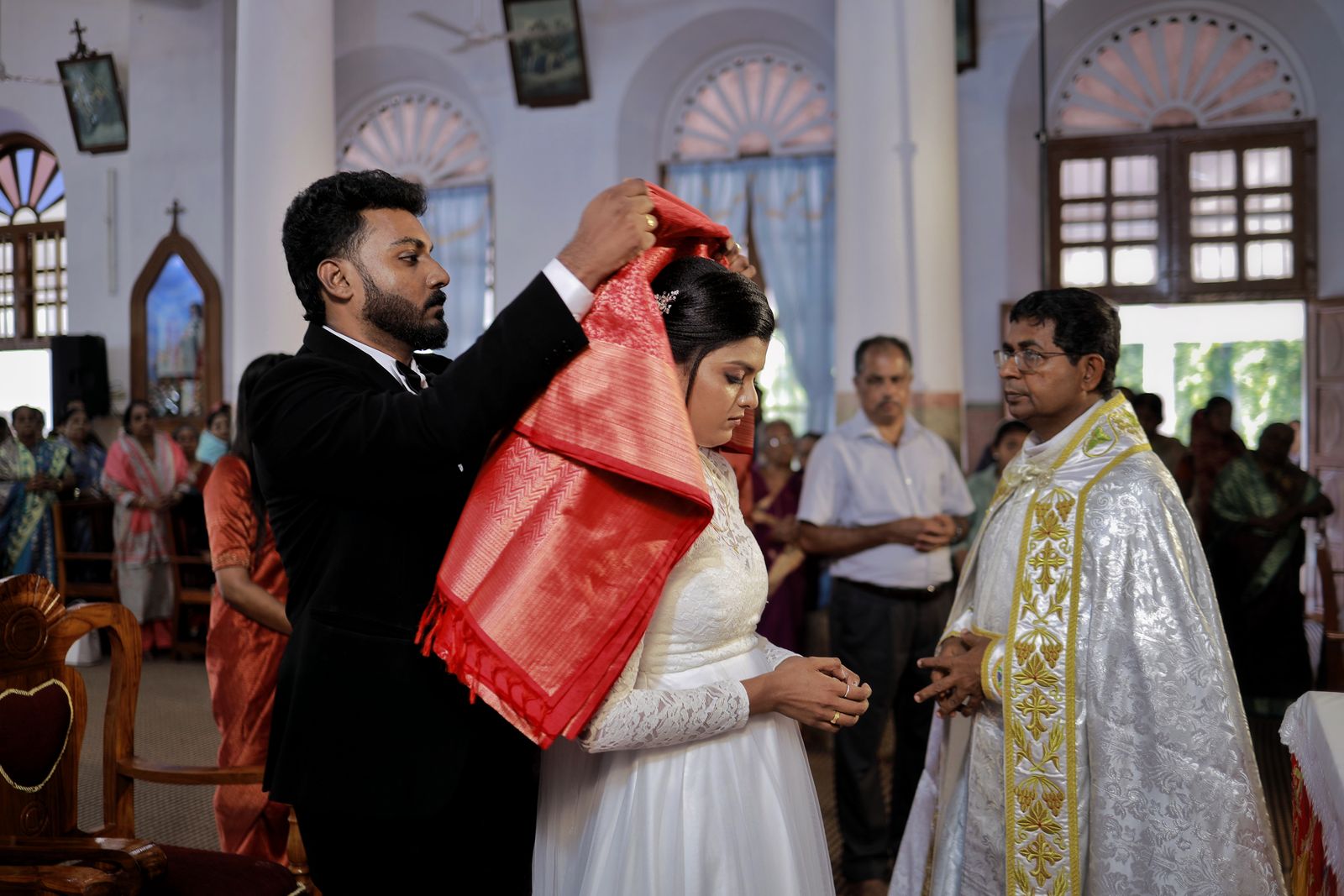 A groom drawing a red silk manthrakodi over the bride's veiled head inside a Kerala church.