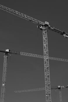 Black and white image of construction cranes towering under a clear sky.