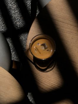 A glass of iced coffee on a wooden table with dramatic shadows, evoking a moody atmosphere.
