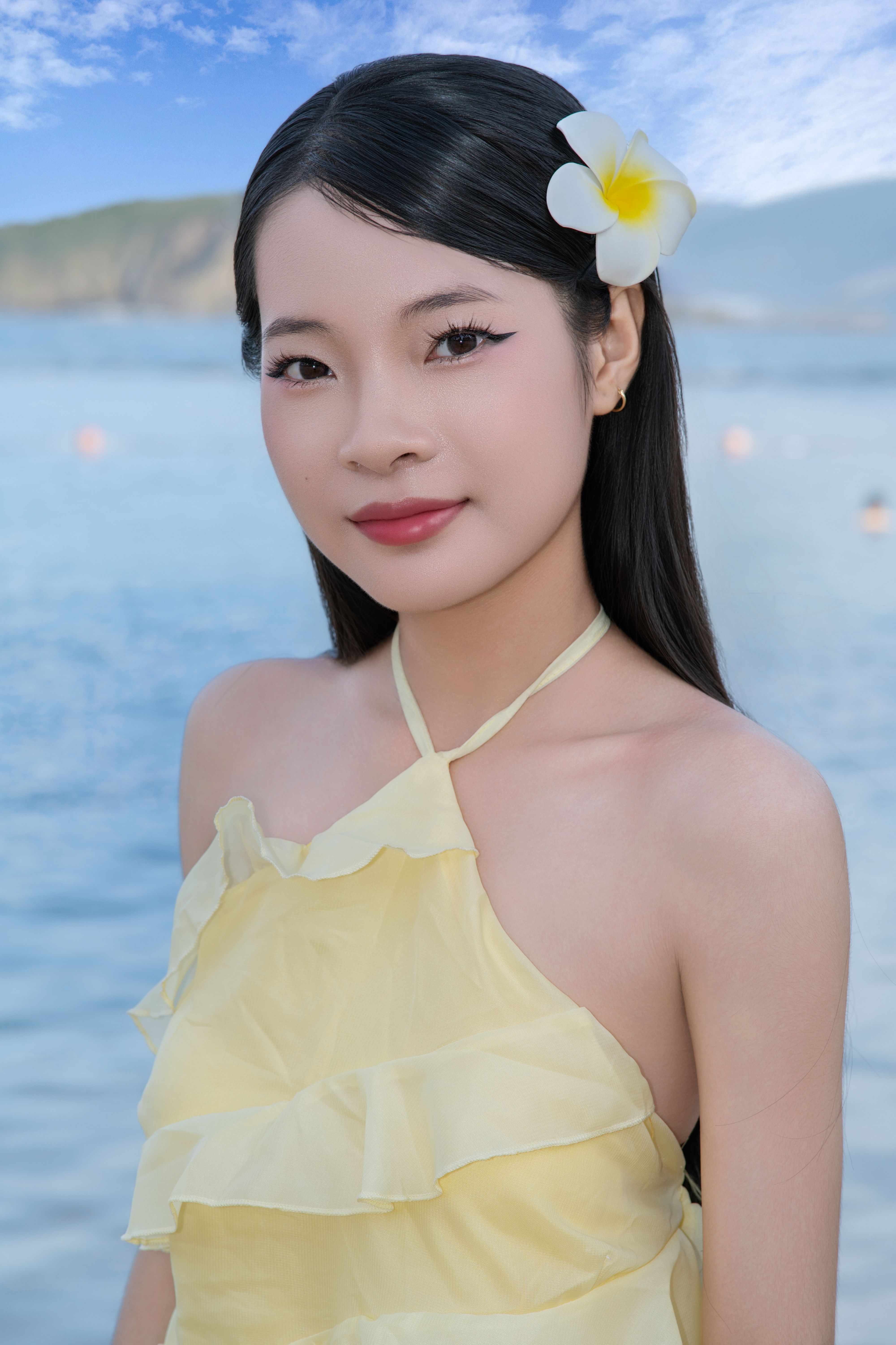 Smiling woman in yellow dress with flower in hair by the beach on a sunny day.