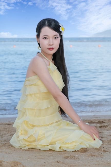 Elegant young woman in a yellow dress, posing on a sandy beach with ocean backdrop.