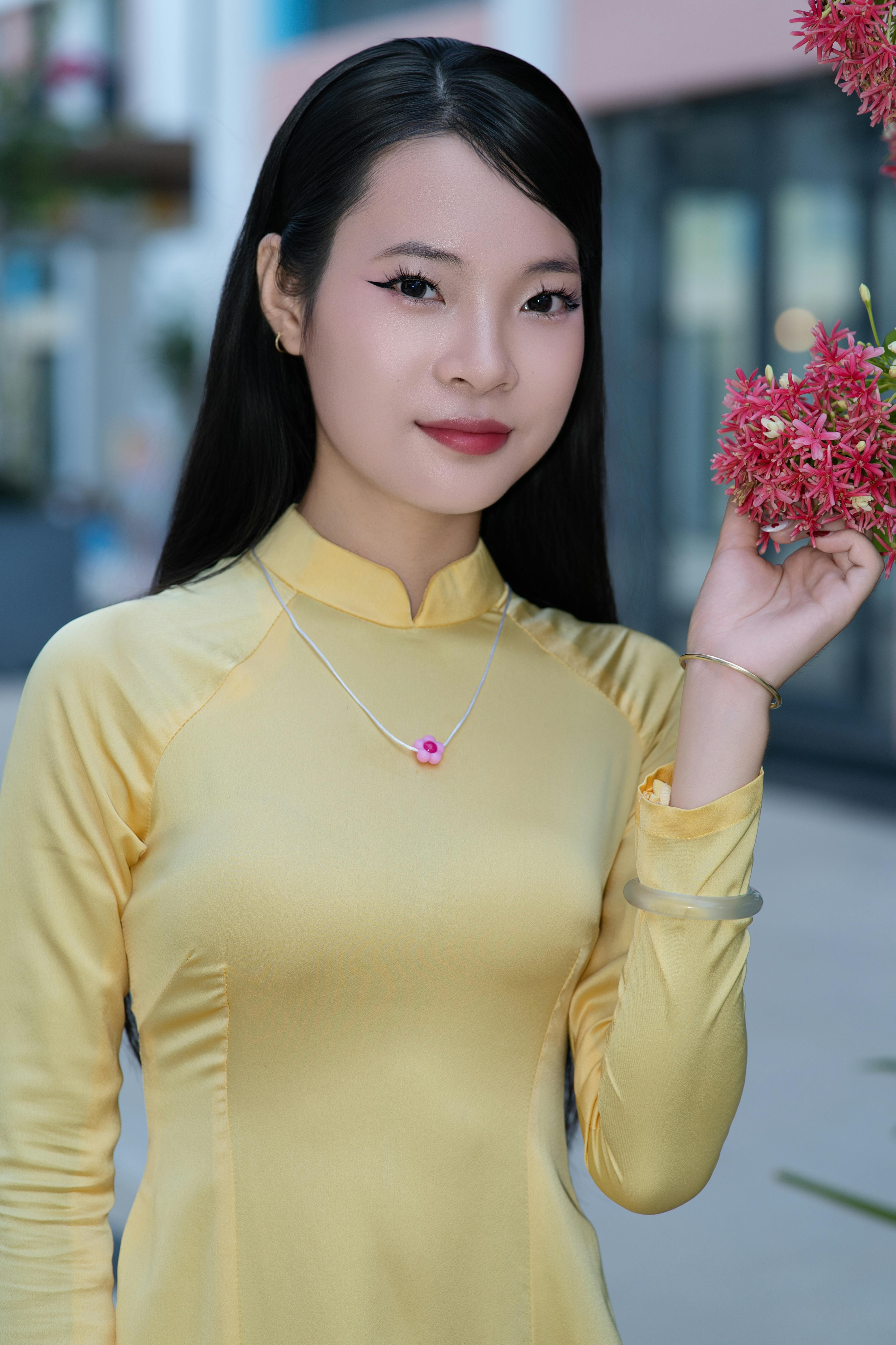 Smiling woman in yellow áo dài holding pink flowers, outdoors.