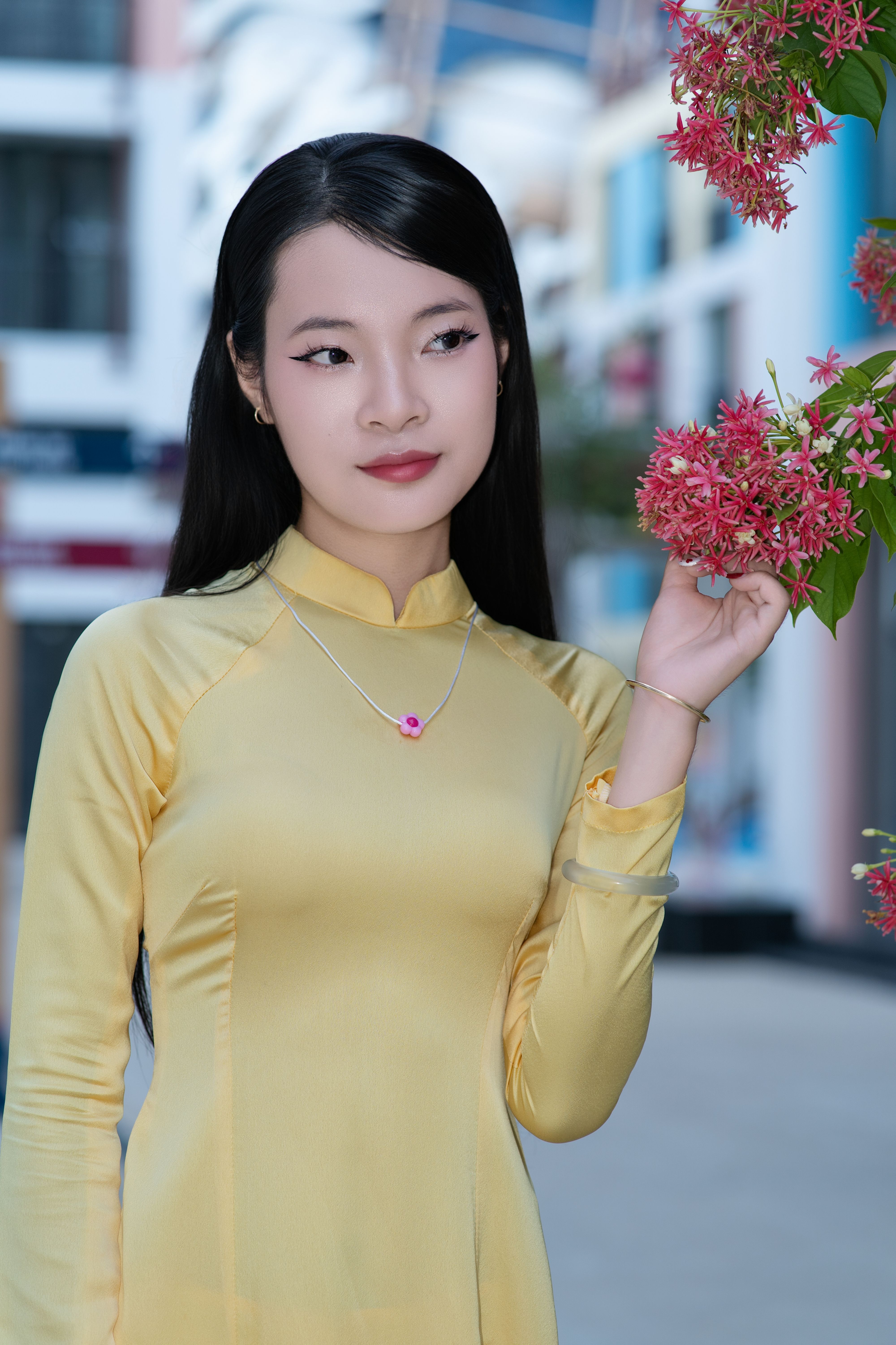 A young woman wearing a yellow Ao Dai admires vibrant flowers outdoors.