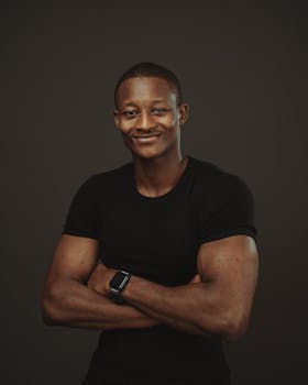 Confident young man in a black t-shirt posing with arms crossed against a dark background in a studio.