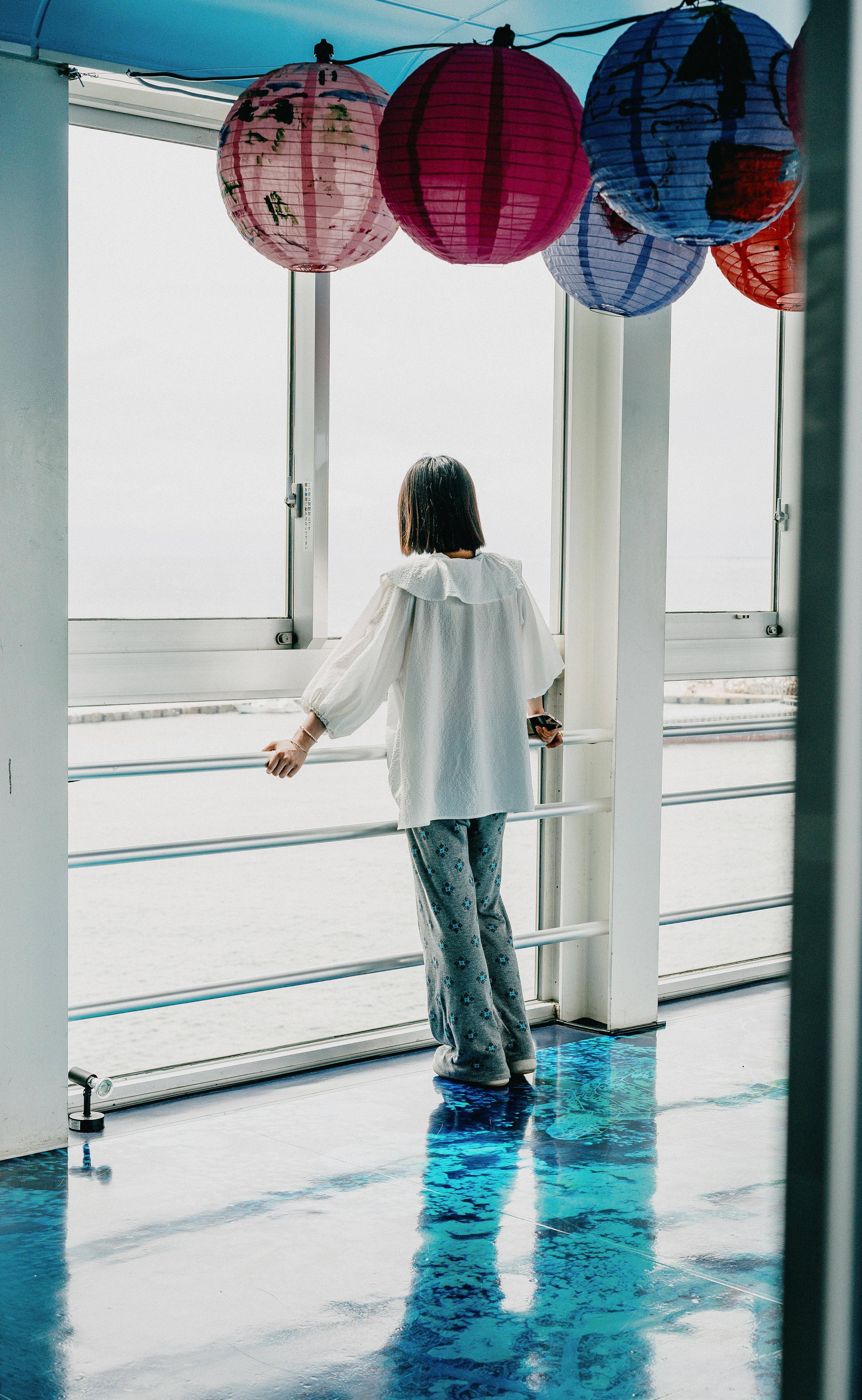 A woman stands indoors, admiring the ocean view, surrounded by vibrant paper lanterns.