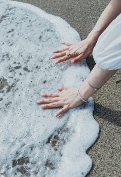 A close-up of hands touching the foamy ocean waves on a sandy beach, capturing a serene moment.