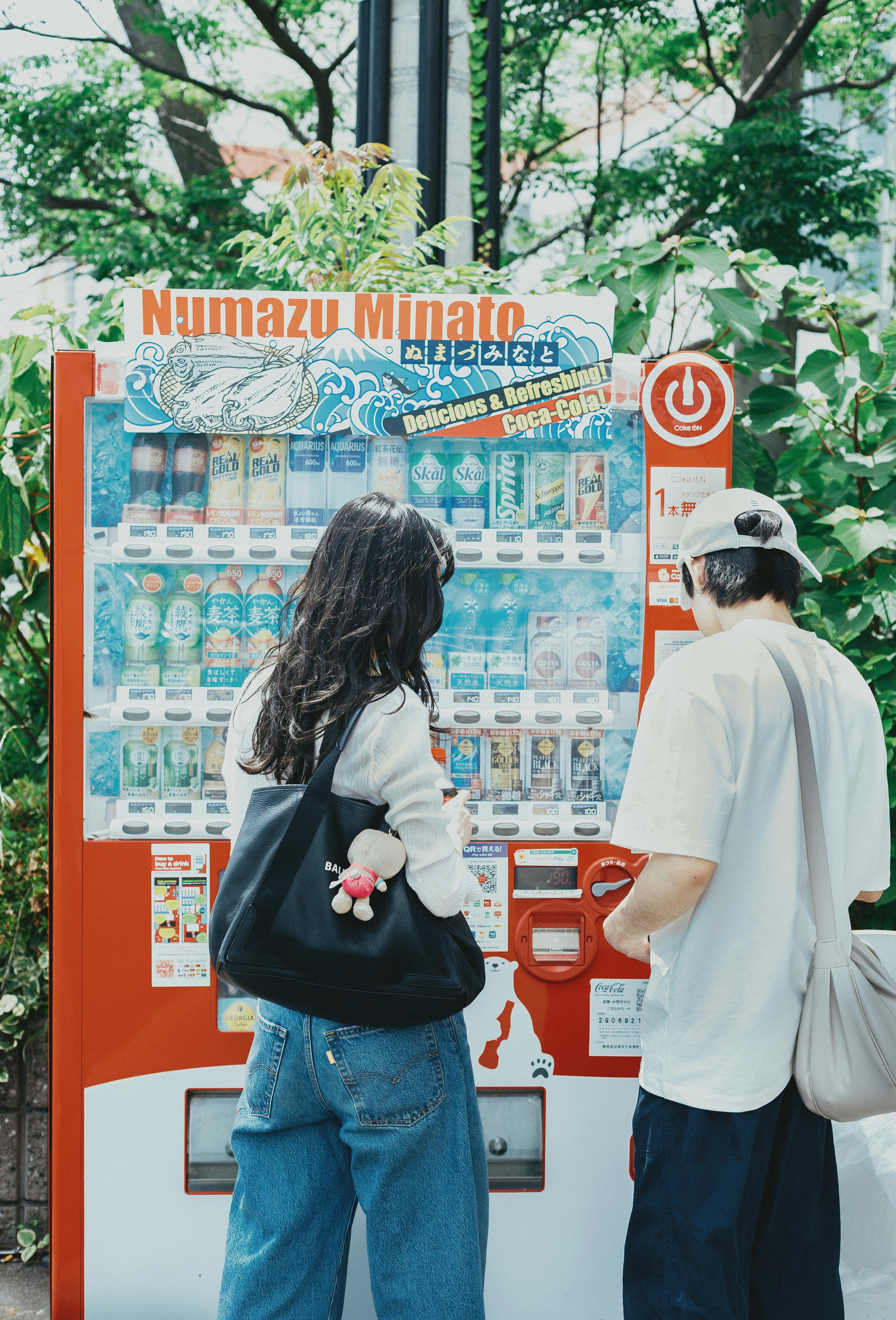 Two individuals at a vending machine in a verdant setting in Numazu, Japan.