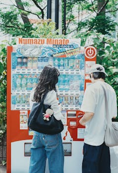 Two individuals at a vending machine in a verdant setting in Numazu, Japan.
