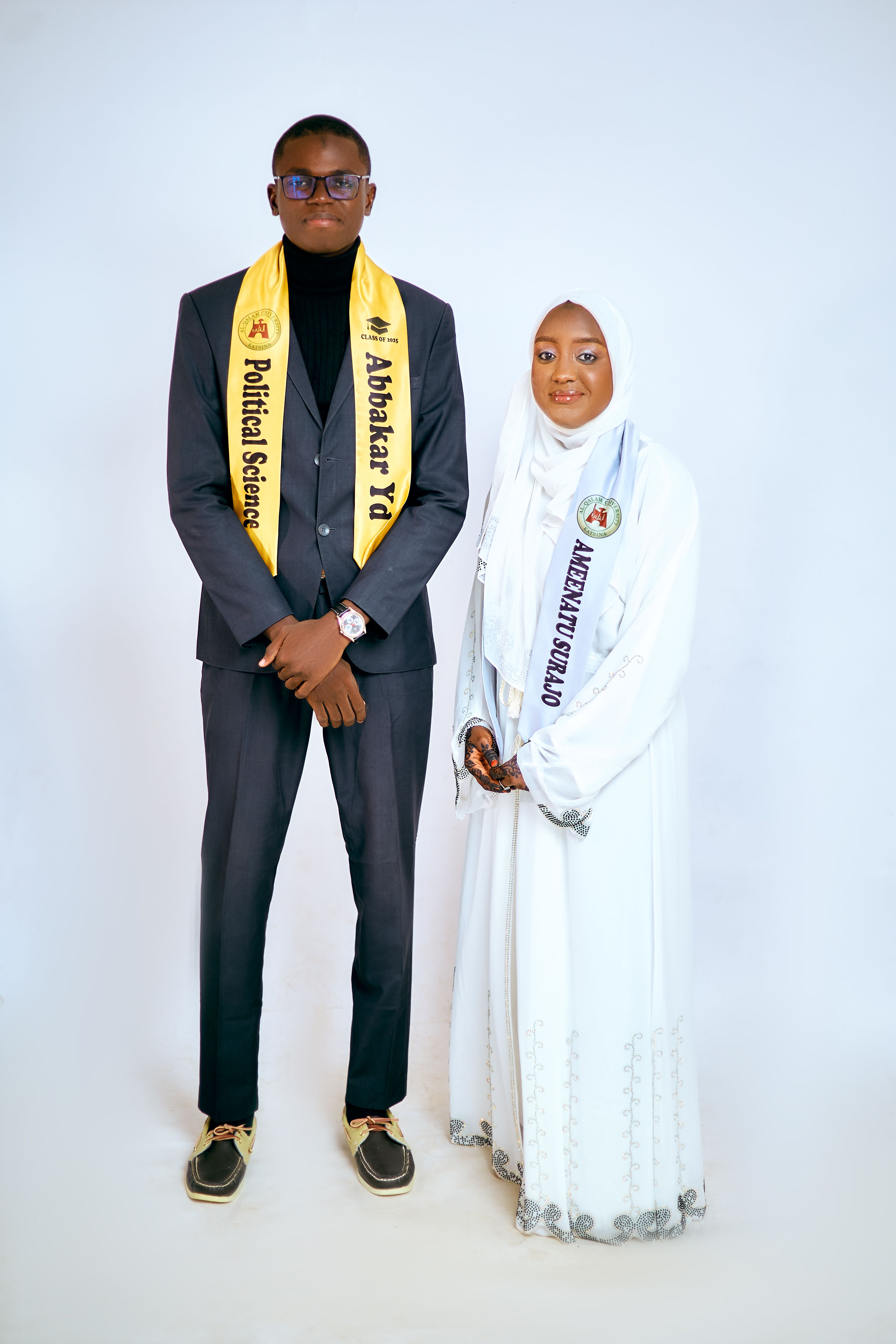 Two graduates posing in formal attire with graduation sashes on a white background.