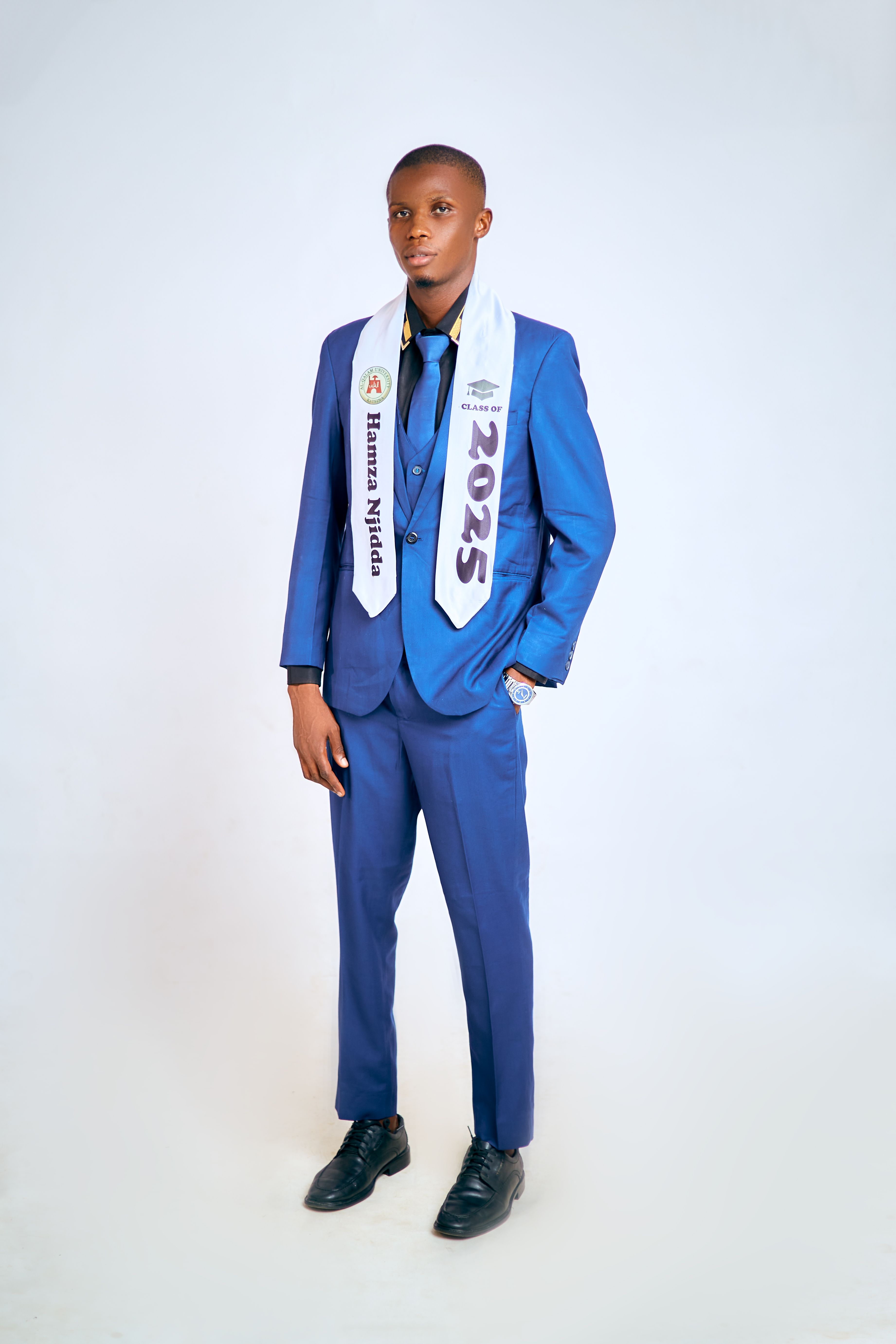 Young adult wearing a 2025 graduation sash, standing in a blue suit against a white background.